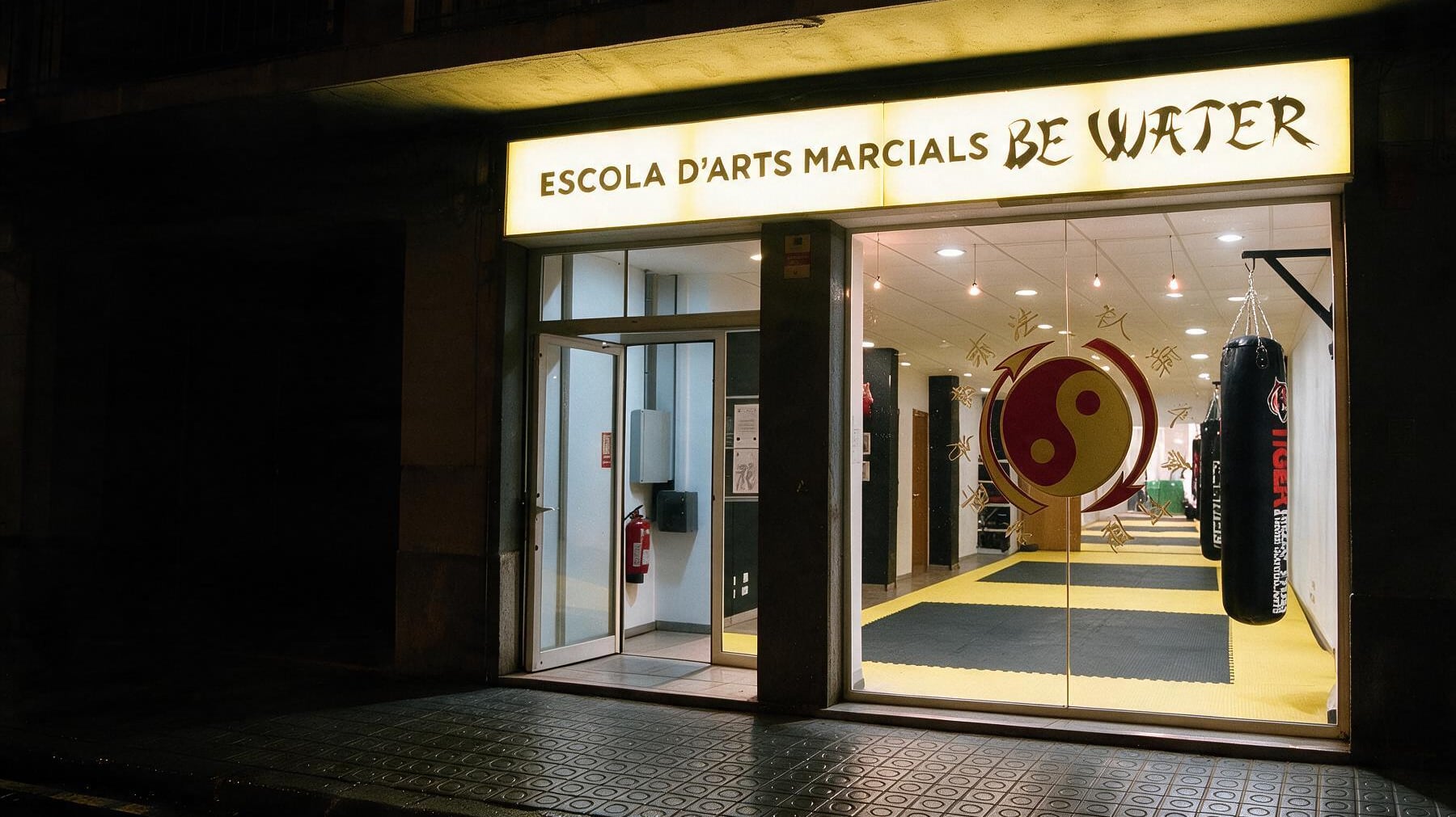 Fachada de noche de la Escola d'Arts Marcials Be Water en Manresa: el rótulo iluminado y, tras el cristal, el tatami, el emblema de Jeet Kune Do y los sacos de boxeo